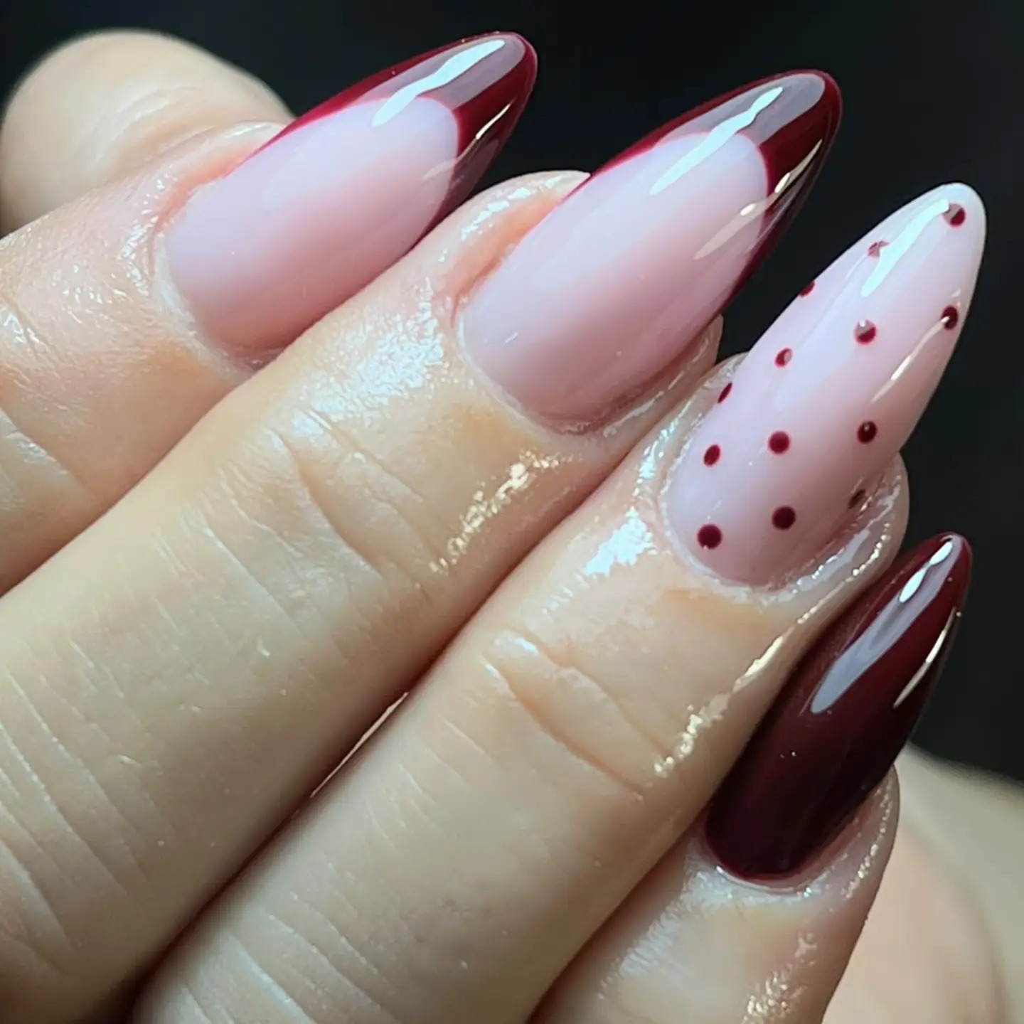 Uñas stiletto largas con degradado vino tinto y una uña con puntos rojos sobre base nude